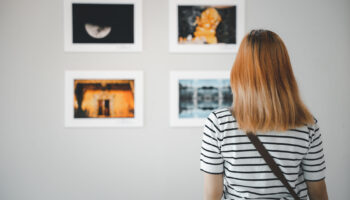 Asian young woman standing she looking art gallery in front of colorful framed paintings pictures on white wall, female watch at photo frame to leaning against at exhibit museum, Back view